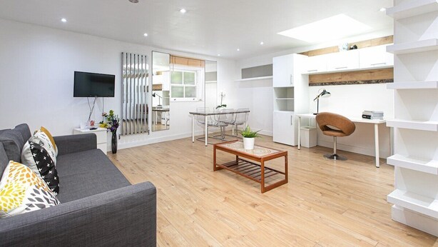 A modern living room with a grey sofa, wooden coffee table, and a TV. There is a dining area with a glass table and chairs, and a kitchen area with white cabinets and a wooden countertop.