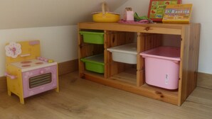 A small wooden playhouse with a pink and yellow toy kitchen, green and pink storage bins, and a toy refrigerator.