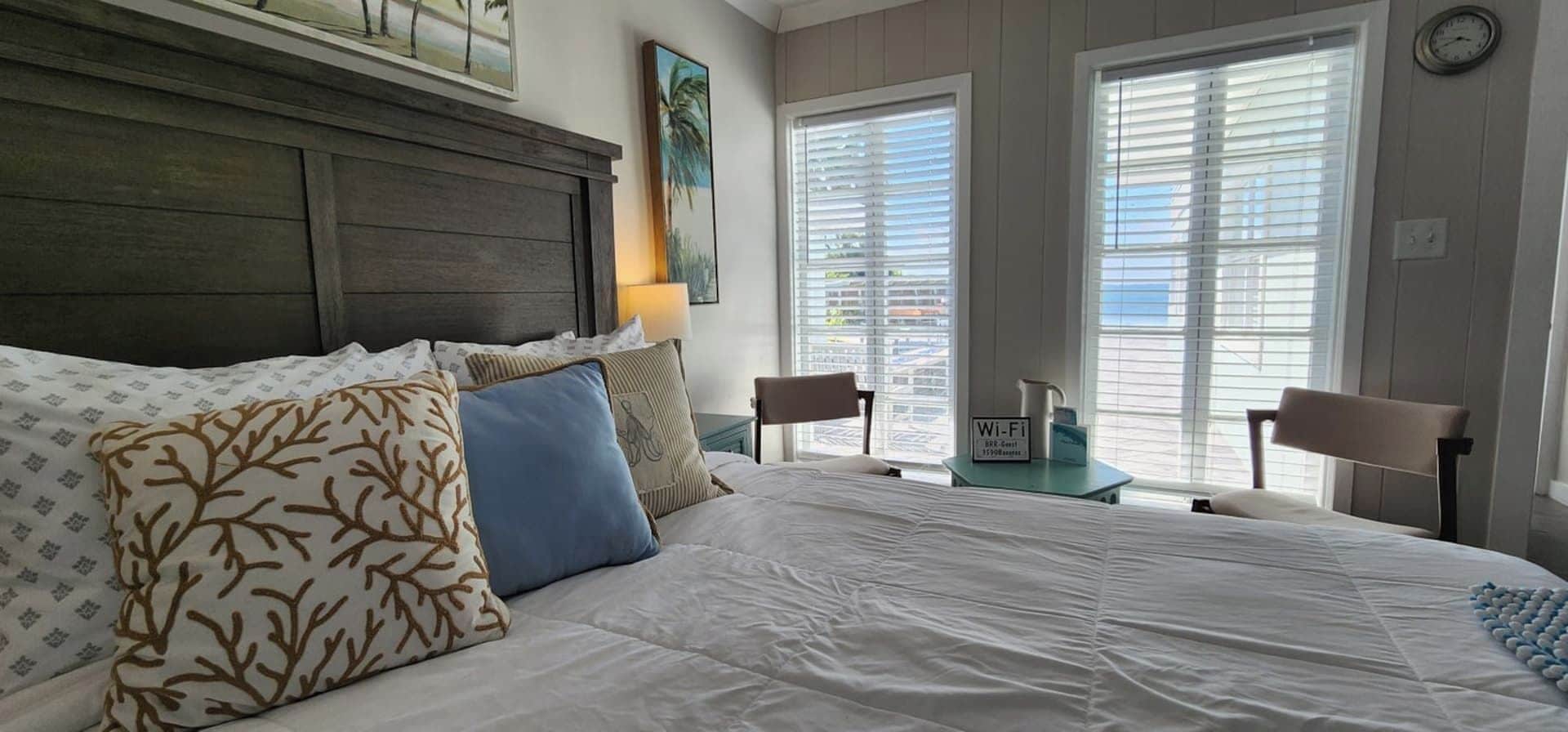 A hotel room with a bed, two nightstands, a lamp, a clock, and a window with blinds.