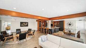 A modern living room with a white sofa, leather chairs, a dining area with wooden chairs, and a kitchen with wooden cabinets and a black refrigerator.