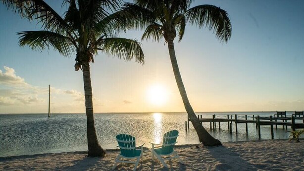 A beach scene with two chairs facing the ocean, palm trees, and a pier. - Compass 2BR Suite - Gourmet Kitchen - Water view (Cocoa Beach)