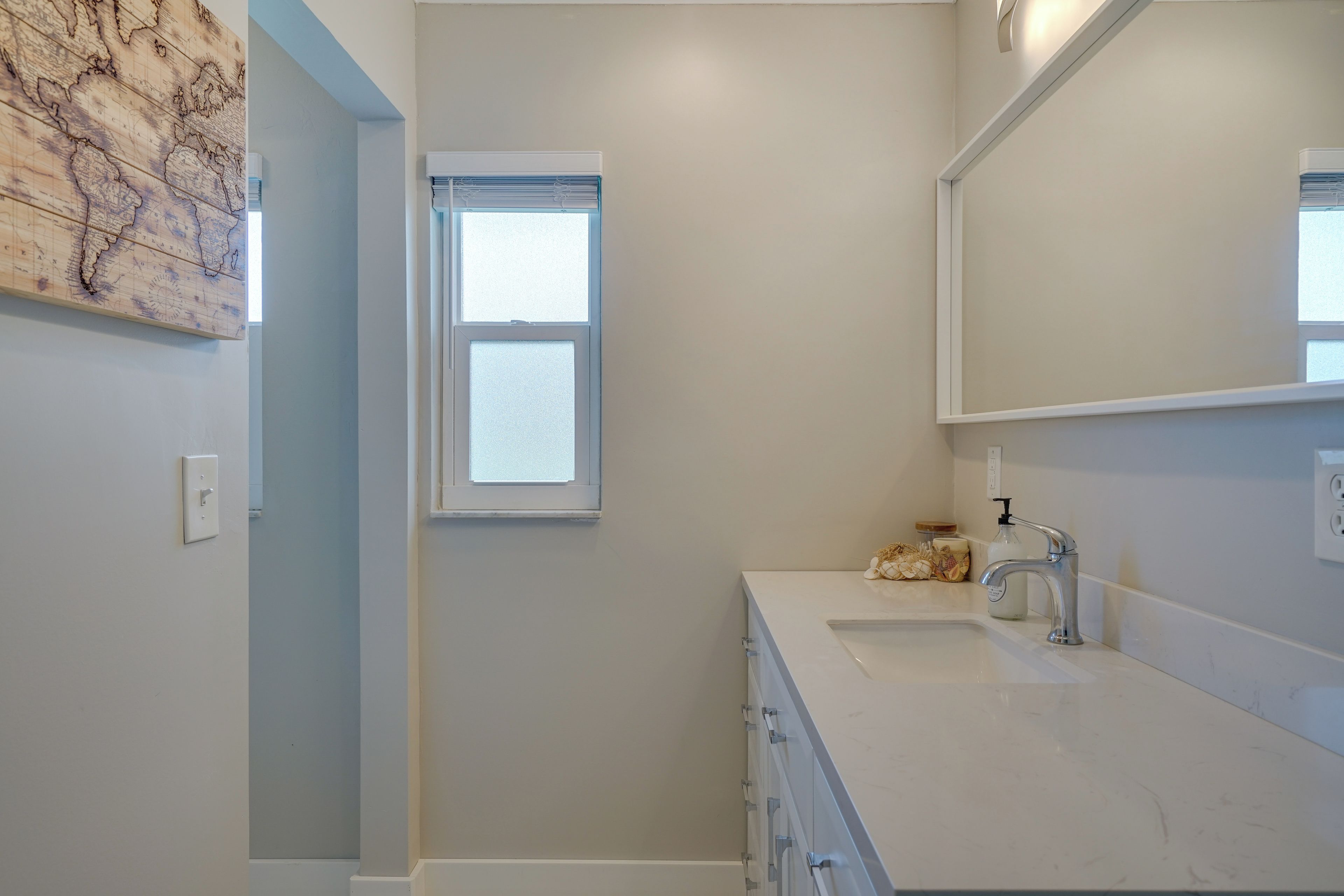 Un baño con tocador blanco, un espejo, una ventana y una lámpara fijada a la pared.