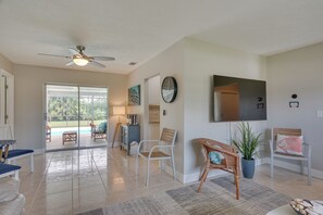 A spacious living area with a ceiling fan, a large flat-screen TV, and a view of a swimming pool through glass doors.