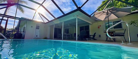 A swimming pool with clear water, surrounded by a covered patio area with chairs and tables.