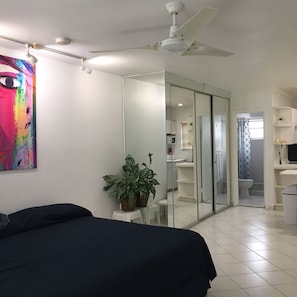 A bedroom with a bed, a ceiling fan, a large mirror, a potted plant, and a bathroom visible through a glass door.