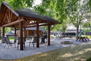 Un espacio al aire libre cubierto con mesas y sillas, equipo de barbacoa, rodeado de árboles y suelo de grava.