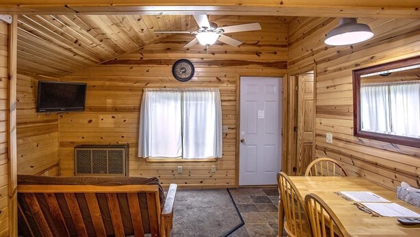 Interior de una cabaña de madera con una mesa de comedor, sillas, un ventilador de techo, un reloj, una puerta y una ventana.