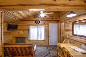 Interior de una cabaña de madera con una mesa de comedor, sillas, un ventilador de techo, un reloj, una puerta y una ventana.