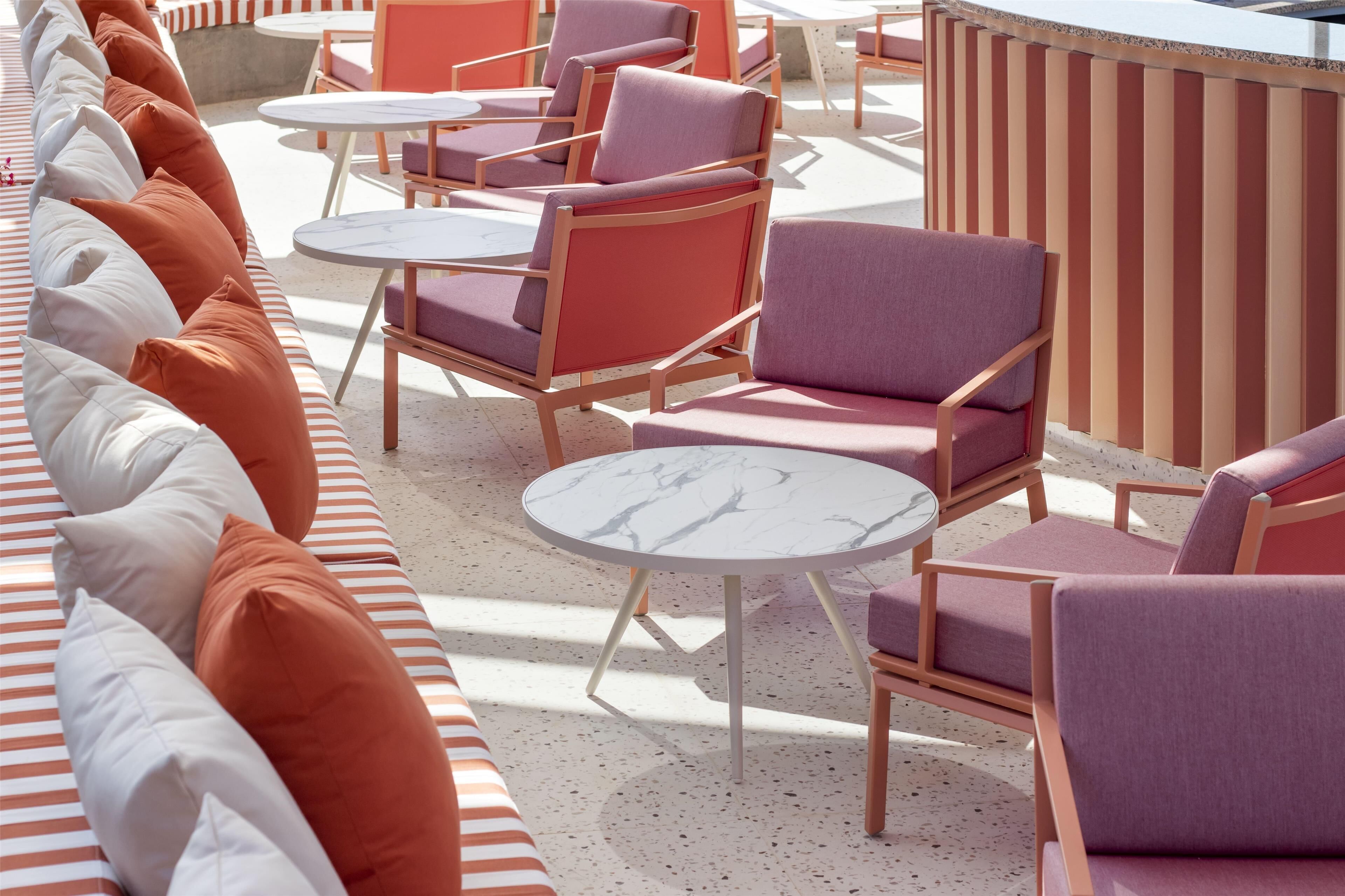 A sunlit outdoor seating area with striped benches and cushioned chairs, featuring a mix of round and square tables.