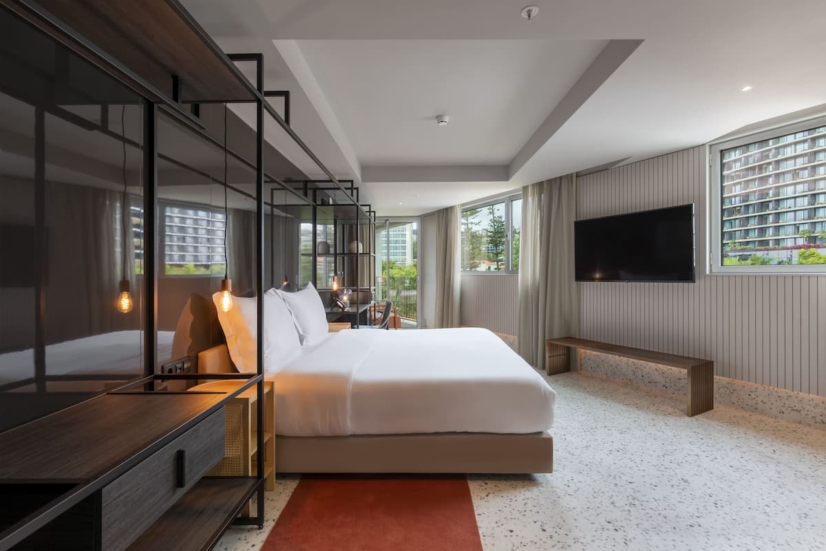 Habitación de hotel moderna con una cama grande, un televisor montado en la pared, un banco de madera y vistas a la ciudad a través de grandes ventanales.