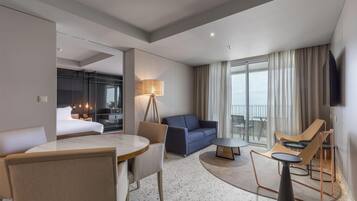 A modern hotel room with a bed, a dining area with a table and chairs, a sofa, a small coffee table, and a TV.