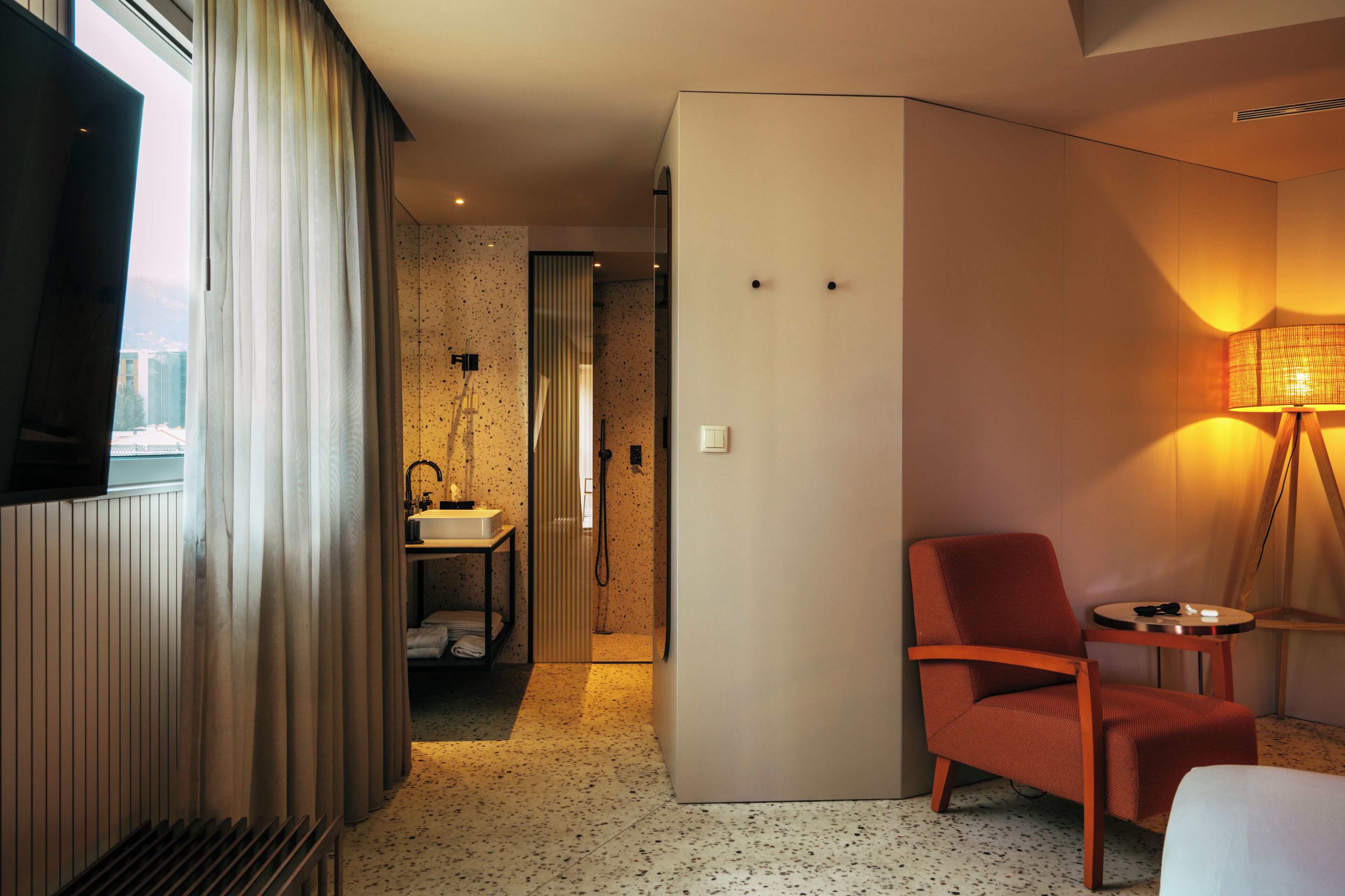 Une chambre d’hôtel comprenant un lit, une chaise, une petite table, une lampe, une salle de bain avec un lavabo et un miroir, et une vue sur la ville par la fenêtre.