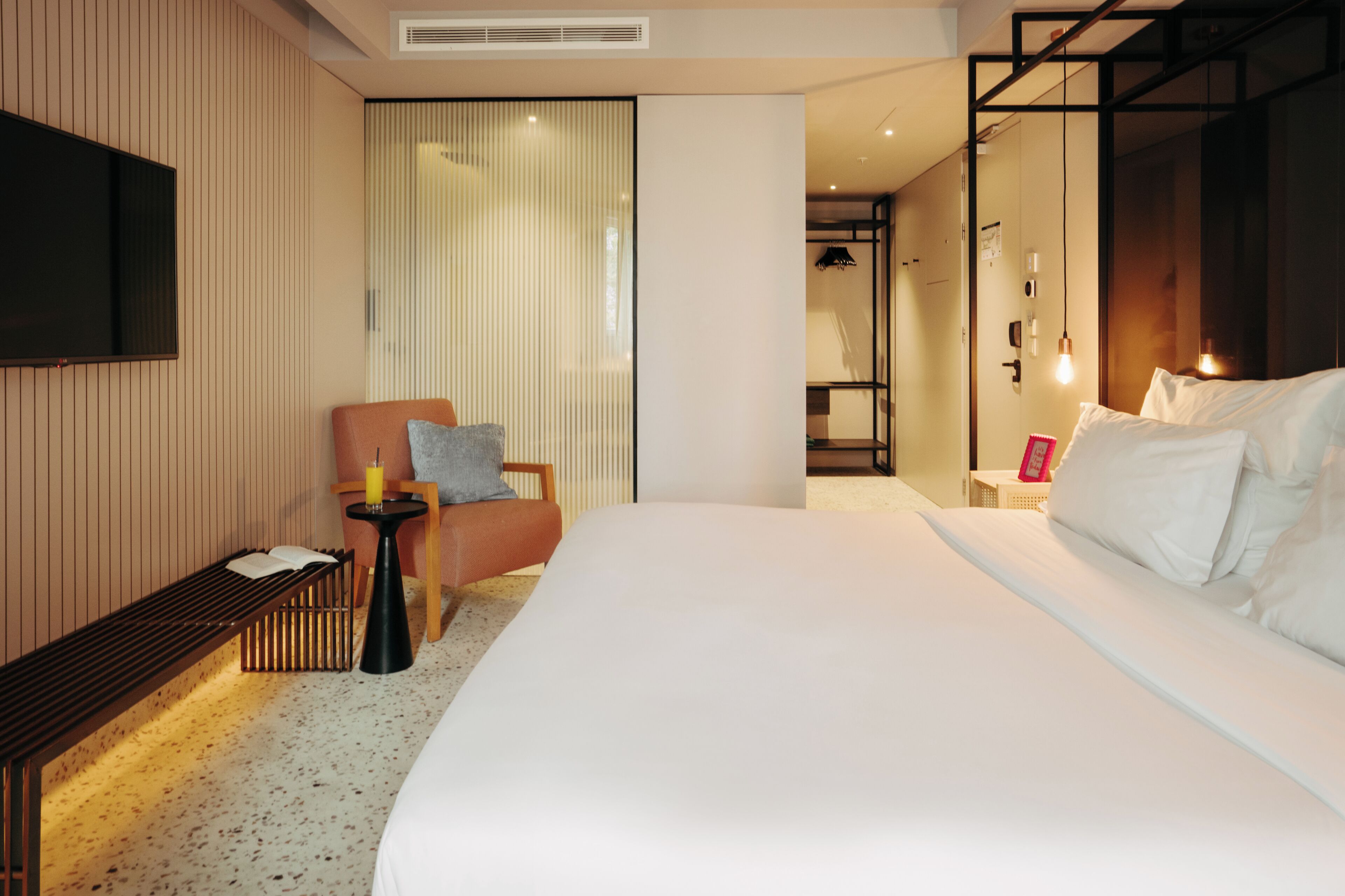 A hotel room with a large bed, a seating area with a chair and a small table, a TV mounted on the wall, and a sliding door leading to a bathroom.