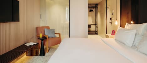 A hotel room with a large bed, a seating area with a chair and a small table, a TV mounted on the wall, and a sliding door leading to a bathroom.