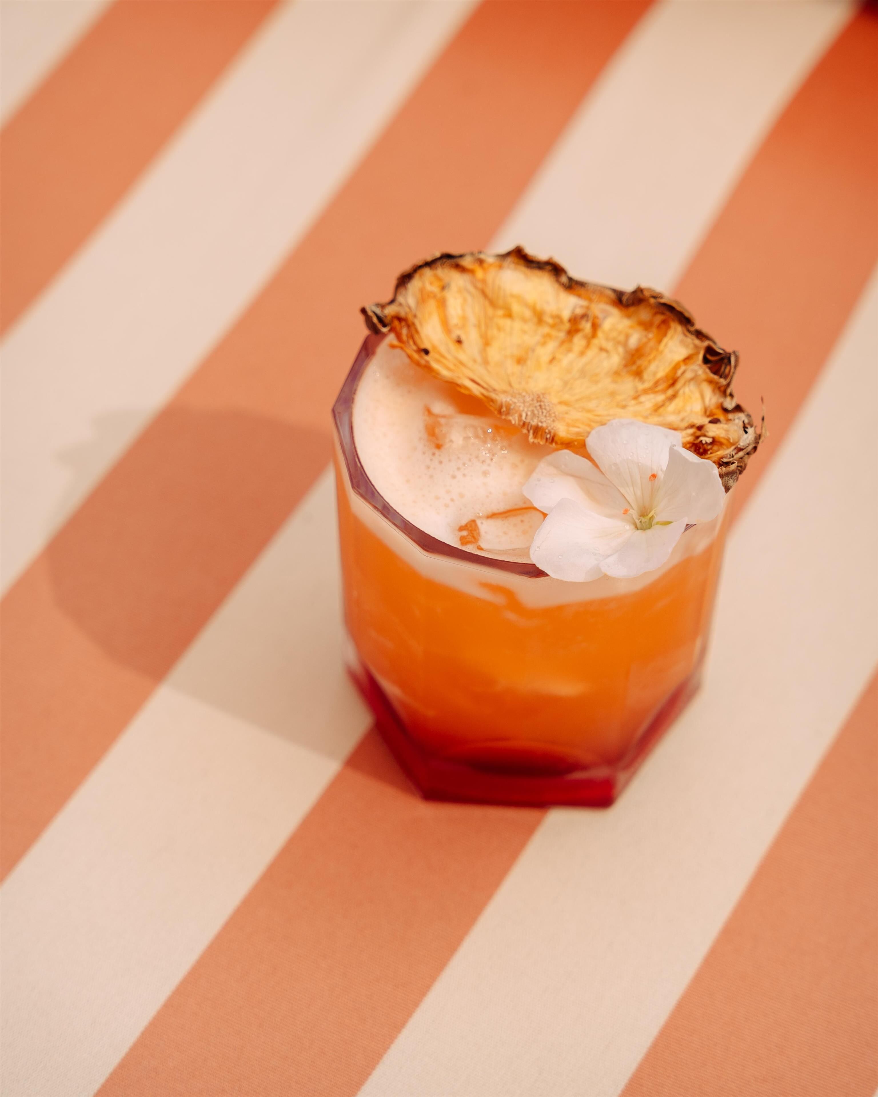 A cocktail glass with a slice of pineapple on the rim, ice cubes, and a flower garnish.