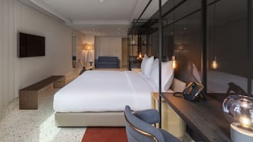 Moderni hotellihuone, jossa on suuri sänky, kirjoituspöytä puhelimineen, tuoli, penkki ja seinälle asennettu televisio.