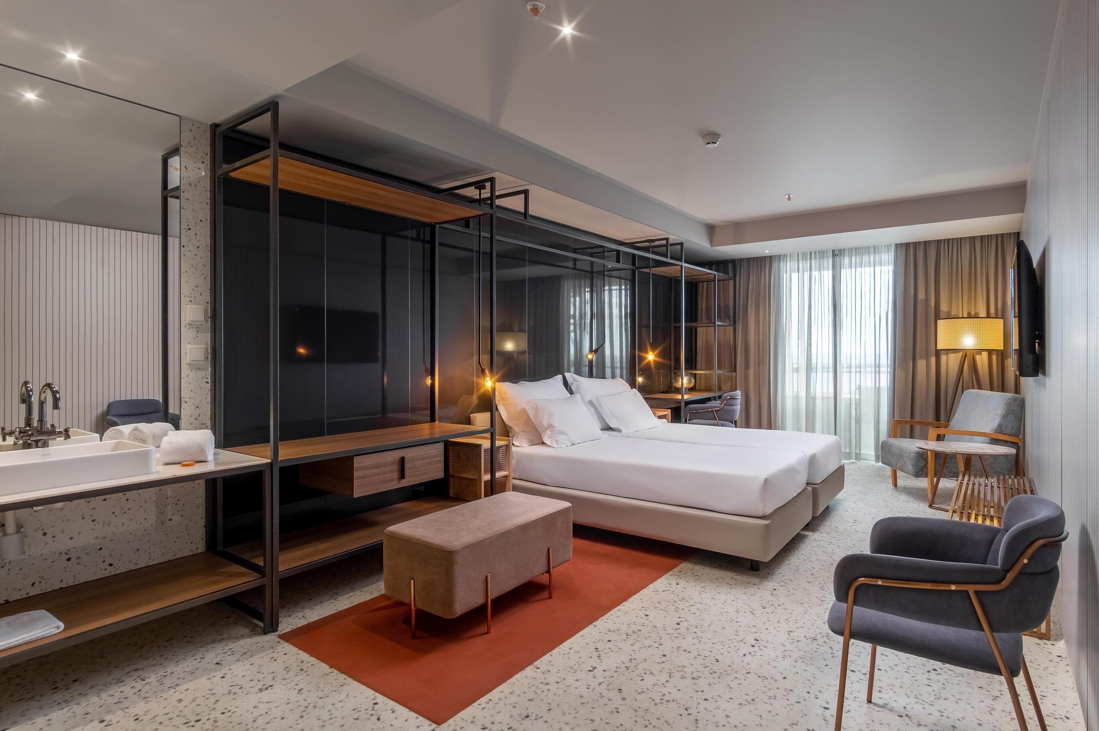 A modern hotel room with a large bed, a desk with a chair, a sofa, a TV, and a bathroom with a sink and mirror.