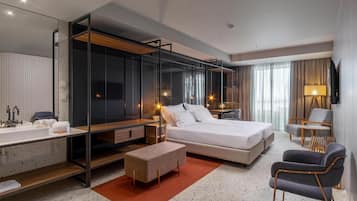 A modern hotel room with a large bed, a desk with a chair, a sofa, a TV, and a bathroom with a sink and mirror.