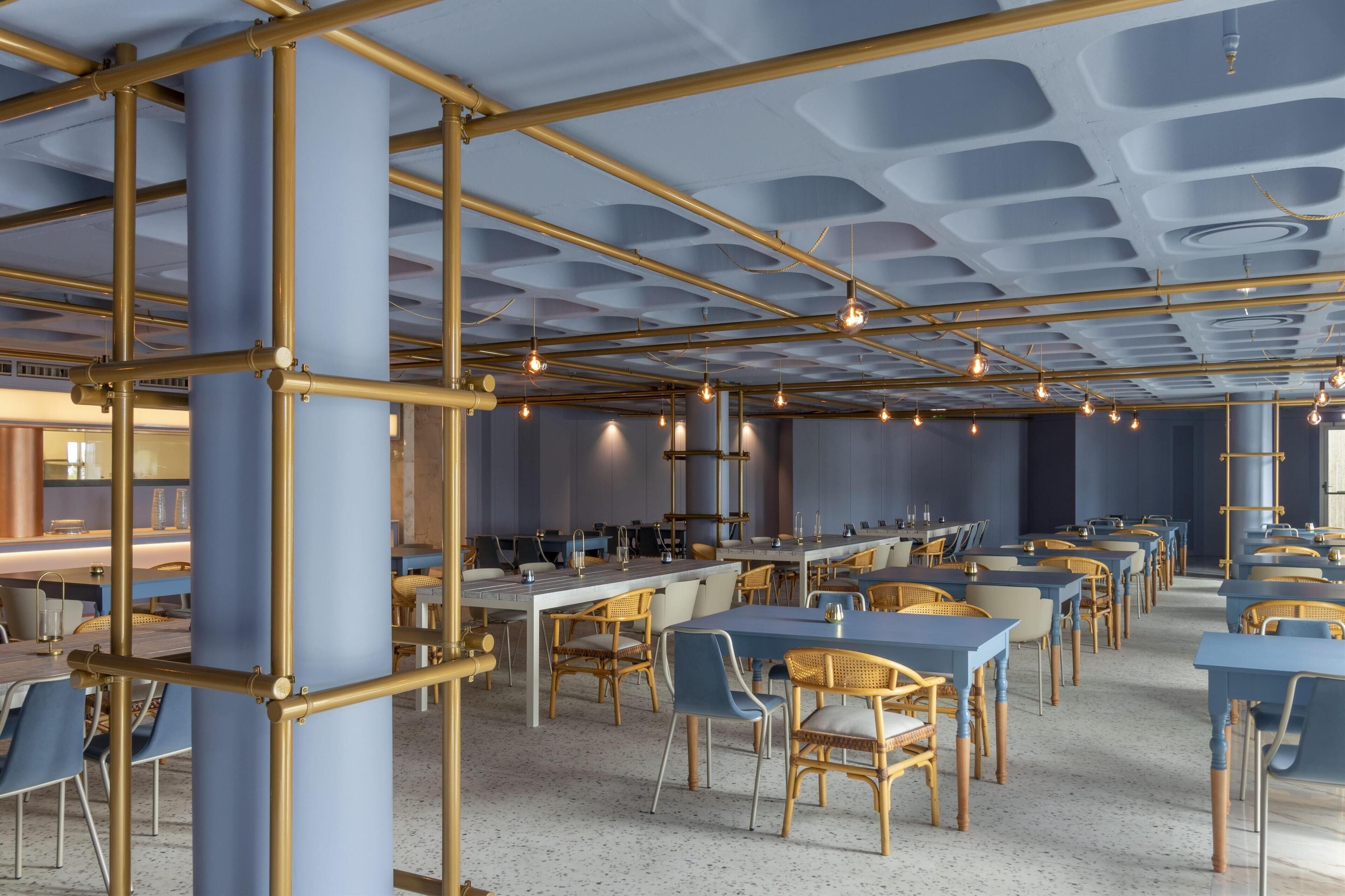 Un vaste espace dédié à la salle à manger, avec des tables et des chaises bleues, des éléments structurels dorés et des luminaires suspendus.