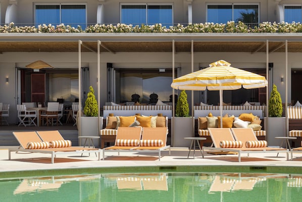 Zona de piscina con sillones de descanso, una sombrilla a rayas y vistas al exterior del edificio.