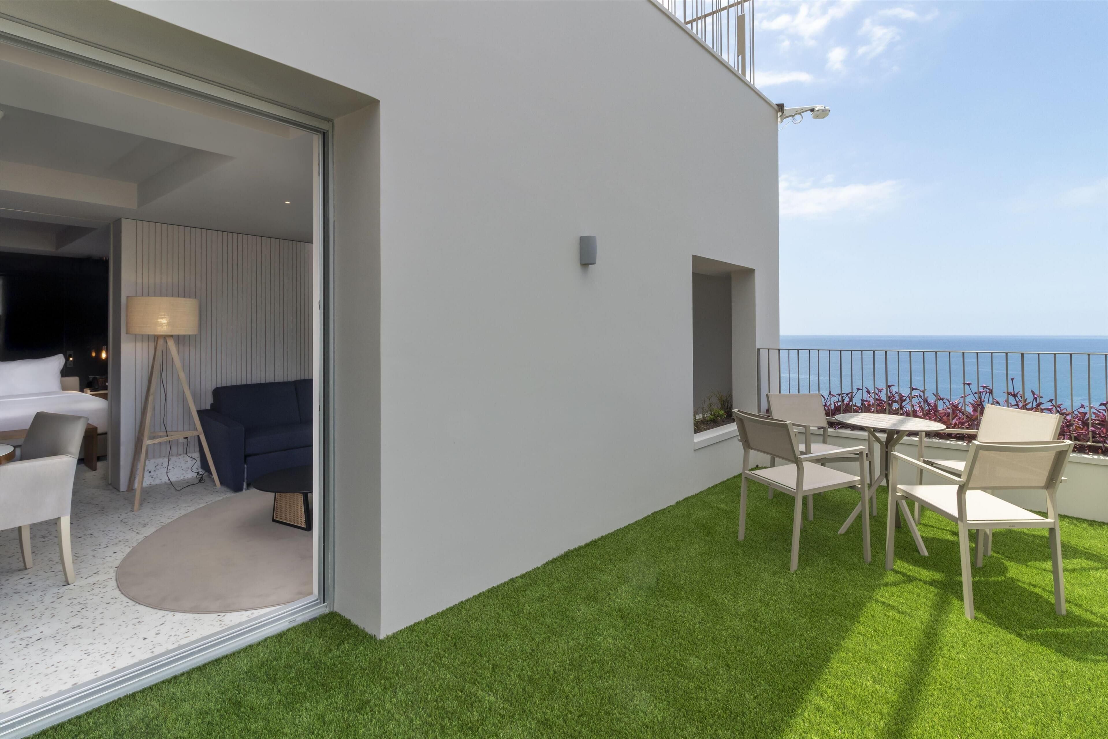 A modern outdoor balcony with a seating area, a view of the sea, and a glass door leading to a room with a bed and a lamp.