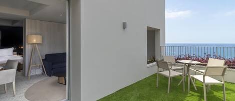 A modern outdoor balcony with a seating area, a view of the sea, and a glass door leading to a room with a bed and a lamp.
