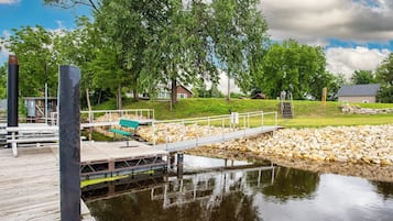 En træbrygge strækker sig ud over stille vand, omgivet af grøn vegetation og en mindre bygning.