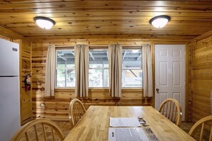 Interior de una cabaña de madera con una mesa de comedor, sillas, un refrigerador y ventanas con cortinas.