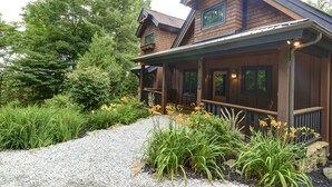 Een houten blokhut met een veranda, omgeven door weelderige begroeiing en een grindpad.