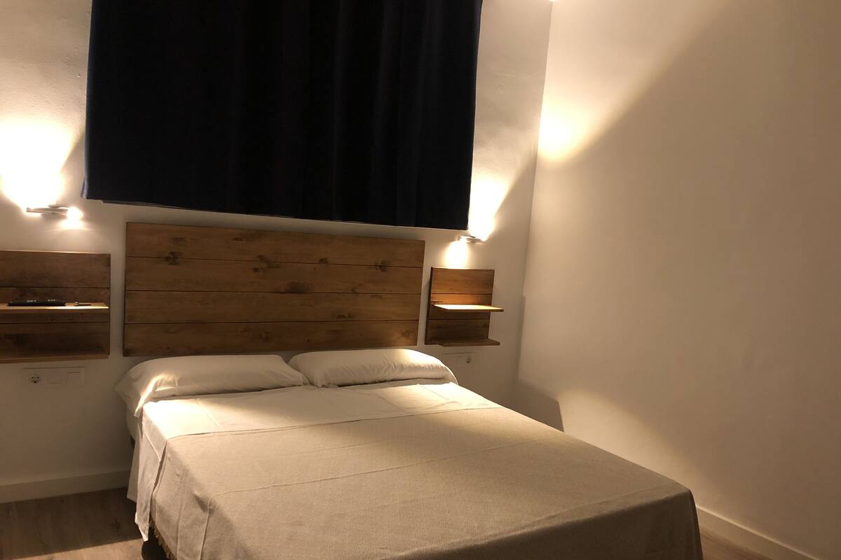 Un dormitorio con cabecera de madera, dos mesitas de noche, cama con colcha color beige y cortina oscura.