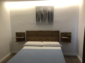 A bedroom with a wooden headboard, a bed with white linens, two bedside tables with small shelves, and a wall-mounted abstract art piece.