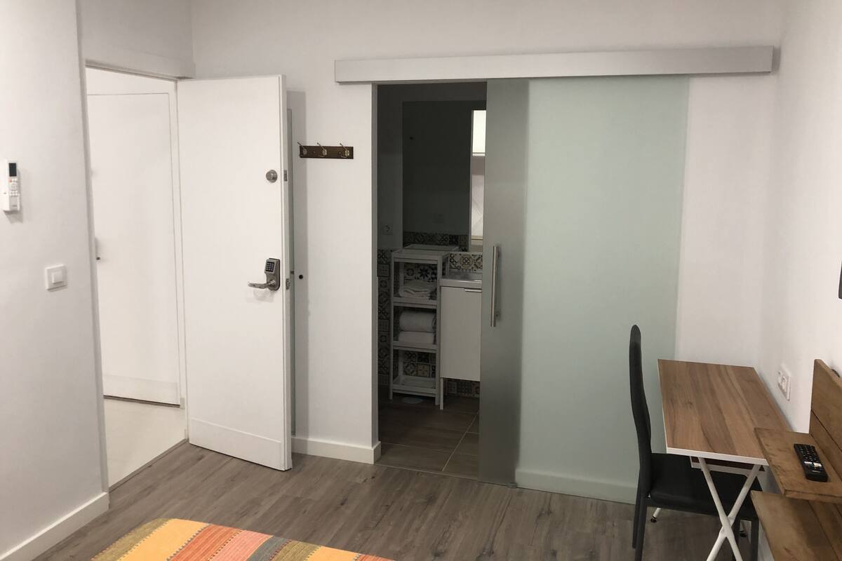 Una habitación con una mesa de comedor de madera, una puerta blanca y una cocina visible a través de un vano abierto.