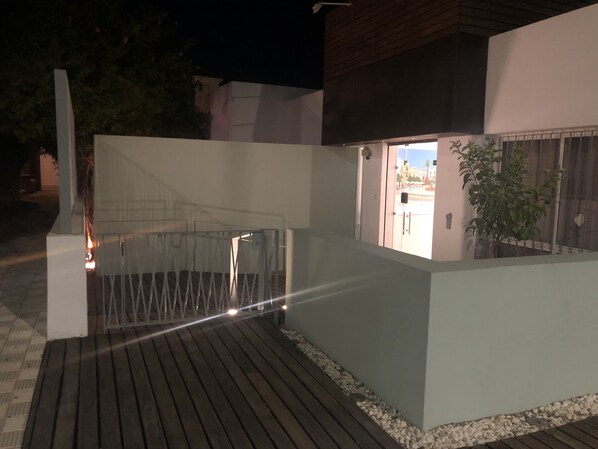 Un espacio exterior moderno con terraza de madera, pared blanca y puerta de metal.