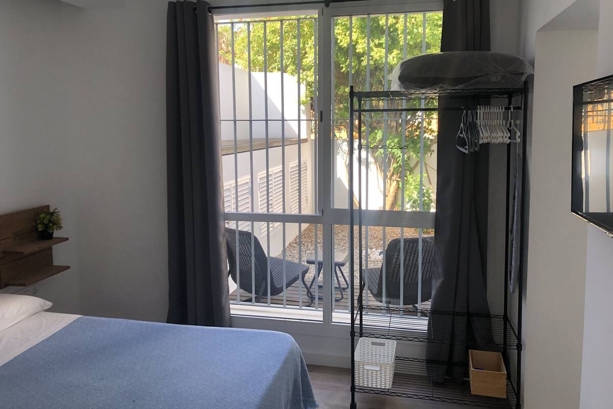Un dormitorio con cama, ventana con cortinas, balcón con una silla y televisión en la pared.