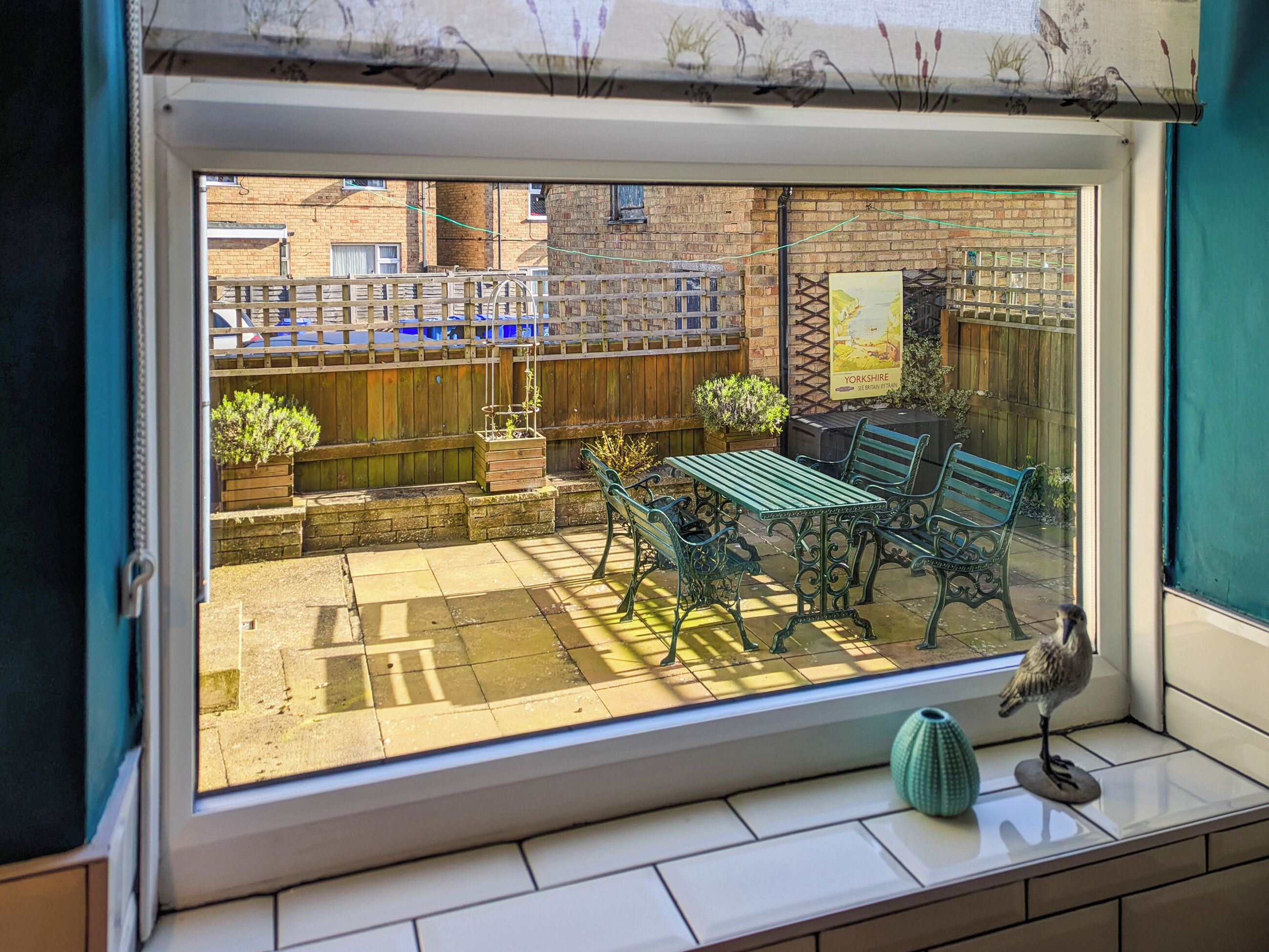 A view through a window of a patio area with a wooden fence, a table, and a bench.