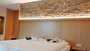 Ein Hotelzimmer mit einer Wand aus Stein, einem Holzkopfteil, einem Bett mit weißer Bettwäsche und zwei gemusterten Kissen sowie einem kleinen Nachttisch aus Holz.