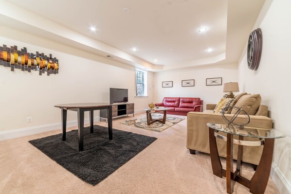 Un salón amplio con un sofá color beige, una mesa de centro de vidrio, una alfombra oscura y un adorno de pared.