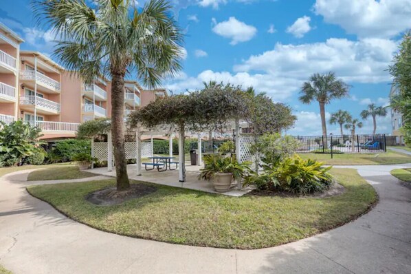 A courtyard with a palm tree, a covered seating area, and a building with balconies. - Flamingo Three (Galveston)