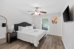A bedroom with a bed, nightstands, a ceiling fan, a window with blinds, and a flat-screen TV.