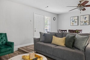 A living room with a grey sofa, a green armchair, a coffee table with a tray and remote, and a ceiling fan. - 30% OFF Arcade Mania - Large Yard 4 pets! 4 TVs (Plano)