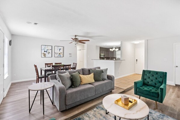 A modern living room with a grey sofa, a round coffee table, and a green armchair. There is a dining area with wooden chairs and a white table. A ceiling fan is visible, and the room has white walls and hardwood flooring.