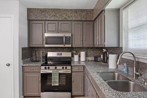 A compact kitchen with a microwave, stove, and sink, featuring gray cabinets and a stone backsplash.
