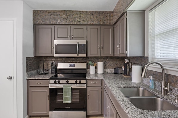 A compact kitchen with a microwave, stove, and sink, featuring gray cabinets and a stone backsplash.
