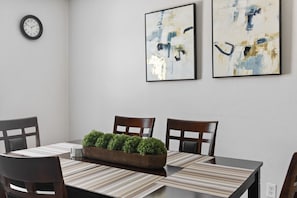 A dining room with a rectangular table, four chairs, a clock, and two abstract paintings on the wall.