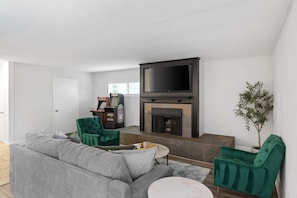 A living room with a grey sofa, green armchairs, a flat-screen TV, a fireplace, and a small table.