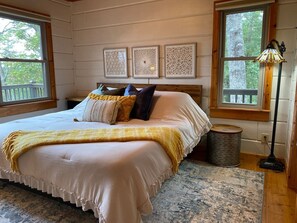 Ein Schlafzimmer mit einem großen Bett, einem Holzkopfende, drei Wandpaneelen zur Dekoration, einem Fenster mit Blick auf Bäume, einer Stehlampe und einem kleinen Papierkorb.