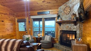 A cozy cabin interior with a leather sofa, two armchairs, a stone fireplace, and a large window overlooking a scenic view. - The Hygge at Blue Ridge - Beautiful Mtn view, 3 Fire types, Hot Tub (Blue Ridge)
