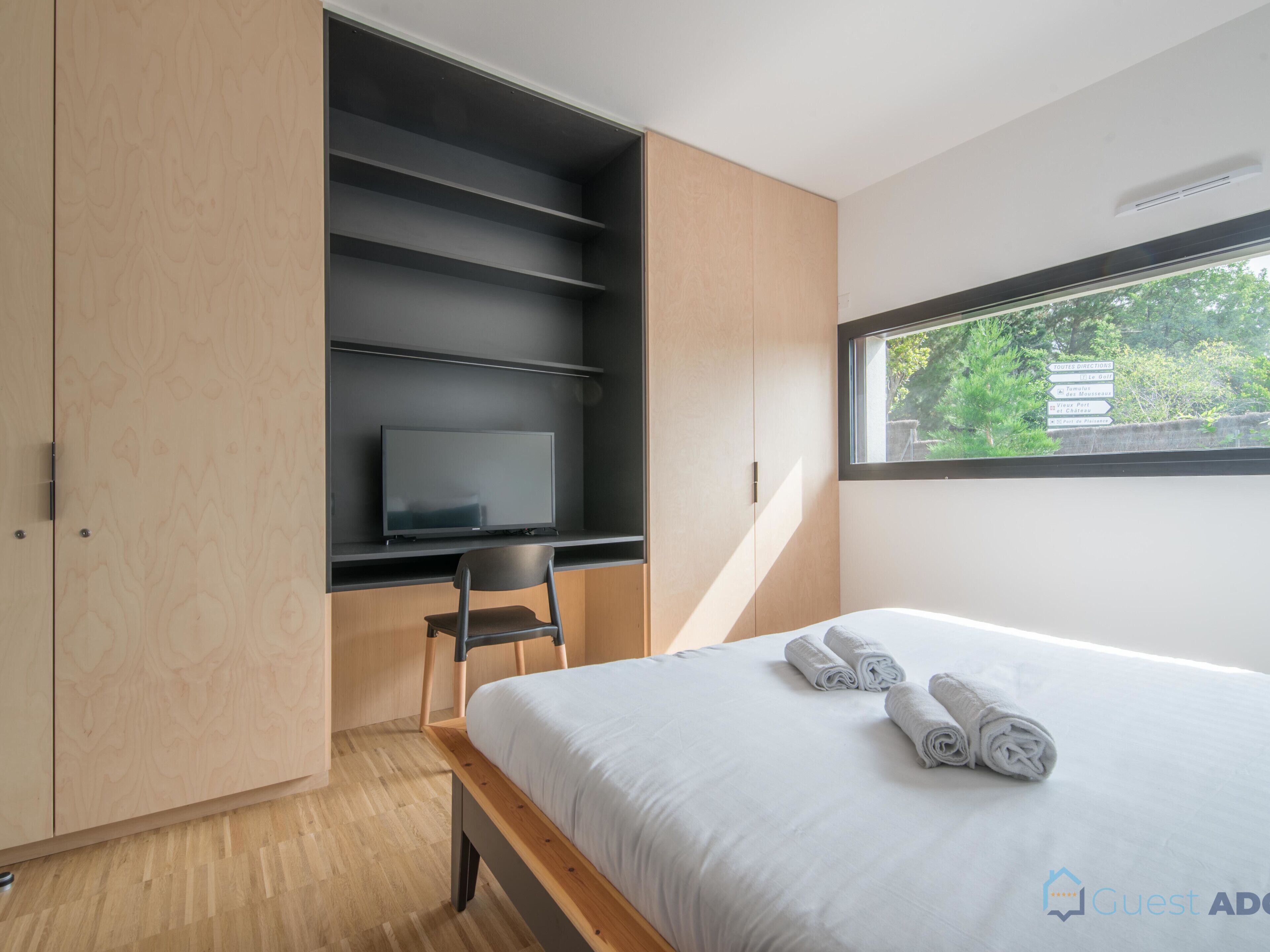 A modern bedroom with a bed, a TV on a shelf, a chair, and a window with a view of greenery.