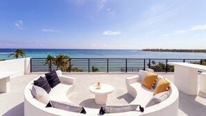 A circular outdoor seating area with a white table and cushions, overlooking a beach and ocean.