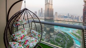 Un fauteuil suspendu orné de coussins à motifs floraux, placé sur un balcon offrant une vue sur un paysage urbain et une étendue d’eau.